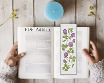 May include: A bookmark with a cross-stitch design of purple flowers and green leaves. The bookmark is placed in an open book. The text "PDF Pattern" is visible on the book's page. A blue candle and dried flowers are on the wooden surface.