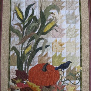 May include: A quilted wall hanging featuring a pumpkin, corn stalks, and fall leaves in shades of brown, green, and yellow. A black bird perches on a vine.