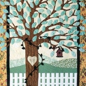 May include: A quilted wall hanging featuring a brown tree with a heart-shaped opening in the trunk. The tree is surrounded by a white picket fence and a green grassy field. A small birdhouse hangs from a branch. The background is a yellow and green floral pattern.