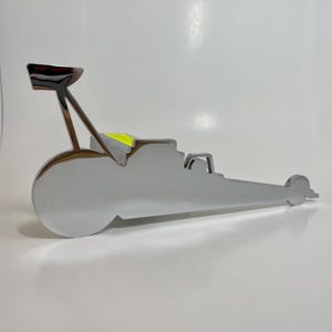 May include: A chrome-plated metal silhouette of a dragster car. The car is in a classic dragster style with a large front wheel and a long, narrow body.