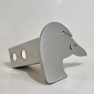 May include: A silver metal hitch cover with a cutout of a goose head. The hitch cover is designed to fit a standard 2-inch receiver hitch.