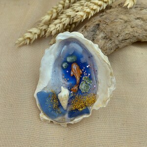 May include: A decorative oyster shell featuring a resin-filled interior with a painted orange koi fish, small green stones, a white seashell, and gold glitter accents. The shell has a smooth, glossy finish and irregular, natural edges.