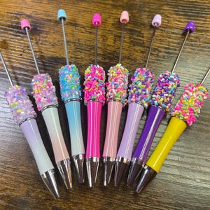 May include: A set of eight pens with colorful, sparkly, sprinkle-like designs. The pens have silver caps and silver tips.