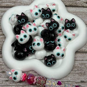May include: A collection of cat-shaped beads in black and white, with large blue eyes and pink accents, are displayed in a white, flower-shaped dish. A beaded bracelet with pink and crystal beads is in the foreground.