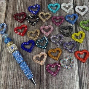 May include: A collection of colorful heart-shaped charms with rhinestones. The charms are made of metal and have a variety of colors, including red, blue, green, and purple.