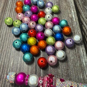 May include: A collection of colorful, iridescent beads in various sizes and hues, including pink, blue, green, and orange. A decorative pen with a pink and silver design, adorned with rhinestones and floral accents, is also present.