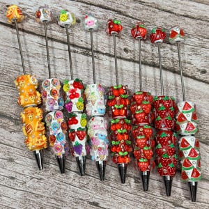 May include: A set of eight metal pens featuring decorative beads with various food-themed designs, including pizza, candy, cherries, ice cream, strawberries, and watermelon slices, arranged in a row on a light wood-grain surface.