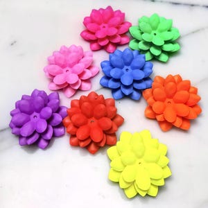 May include: A collection of eight multi-layered flower-shaped beads in vibrant neon colors including pink, green, blue, orange, purple, red, and yellow. Each bead features a central hole and rounded petals arranged in a circular, stacked pattern.