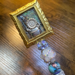 May include: A gold-toned picture frame with a iridescent abalone shell background and a silver flower design. The frame is attached to a beaded charm with white and blue beads.