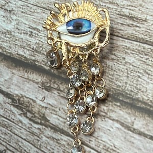 May include: Gold-toned eye-shaped brooch with a blue iris and white sclera. The eye is surrounded by a gold sunburst design. Clear, dangling rhinestones hang from the bottom of the brooch, adding sparkle. The brooch is set against a wooden background.