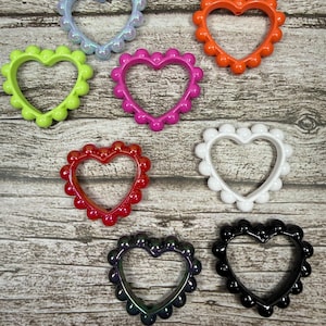 May include: A collection of heart-shaped embellishments in various colors, including green, pink, orange, red, white, iridescent, black, and purple. Each heart has a scalloped edge. The hearts are arranged on a wooden surface.