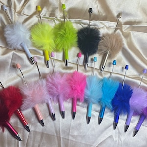May include: A collection of 18 colorful fluffy pens with different colored pom-poms on the top. The pens are arranged in a semi-circle on a gold fabric background.