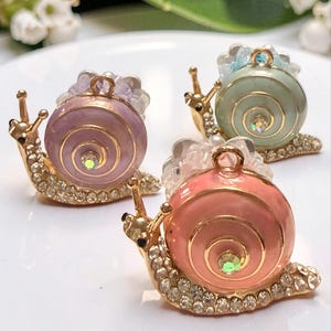 May include: Three snail-shaped charms featuring gold-tone metal bodies encrusted with clear rhinestones. Each snail has a smooth, pastel-colored enamel shell in shades of lavender, mint green, and peach, topped with a small metal loop for hanging.