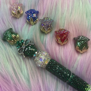 May include: A green pen with a bedazzled barrel and a rose-shaped top. Six rose-shaped embellishments in pink, blue, purple, red, and green are scattered around the pen. The pen and embellishments have gold accents.