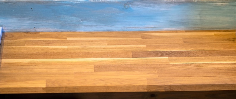 K&ouml;nnte beinhalten: Nahaufnahme einer h&ouml;lzernen Metzgerblock-Arbeitsplatte mit hellbraunem Finish. Die Arbeitsplatte besteht aus rechteckigen Holzplanken. Der Hintergrund ist eine hellblau lackierte Holzoberfl&auml;che.