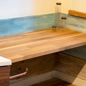 K&ouml;nnte beinhalten: Eine Holz-Arbeitsplatte mit hellbraunem Finish und Kupferrohrdetails. Die Arbeitsplatte befindet sich vor einer blau und wei&szlig; gestrichenen Wand. Klare Glasflaschen mit dunklen Deckeln stehen darauf.