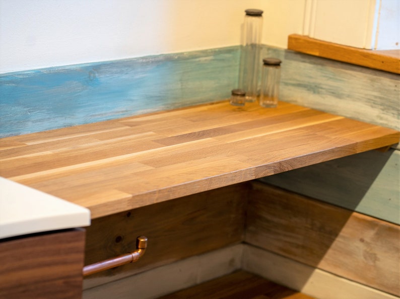 K&ouml;nnte beinhalten: Eine Holz-Arbeitsplatte mit hellbraunem Finish und Kupferrohrdetails. Die Arbeitsplatte befindet sich vor einer blau und wei&szlig; gestrichenen Wand. Klare Glasflaschen mit dunklen Deckeln stehen darauf.