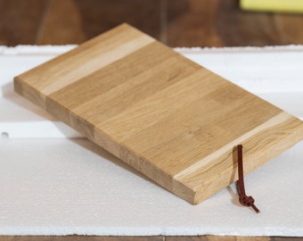 May include: A rectangular wooden cutting board with a light brown finish and a leather strap. The board has a natural wood grain pattern and is made of multiple pieces of wood.