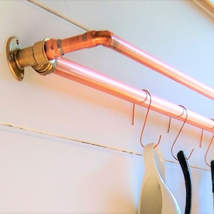 May include: Two copper pipes mounted on a white wall with a brass fitting. The pipes have several hooks attached to them. A white ceramic mug and a black utensil are hanging from the hooks.