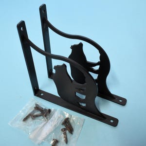 May include: Two black metal shelf brackets with a bear silhouette design. Each bracket has a flat base with screw holes and a vertical support. A small bag of screws is included.