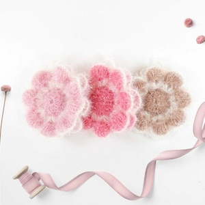 May include: Three crocheted flower-shaped scrubbers in pink and brown shades. The scrubbers are arranged in a row on a white background. A spool of pink ribbon and dried flowers are also visible.