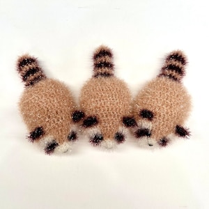 May include: Three crocheted raccoon-shaped ornaments with brown and white fur. The ornaments are arranged in a row, facing the viewer.