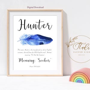 Op de afbeelding: Een ingelijste print met de naam "Hunter" in elegant zwart schrift, boven een blauwe aquarelveer. De tekst eronder legt de betekenis van de naam, "Seeker", uit. Een houten regenboog speelgoed staat ervoor.