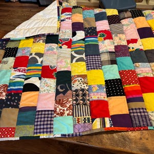 Puede incluir: Un edredón de retazos con una variedad de cuadrados de tela coloridos. El edredón presenta una mezcla de patrones y texturas, incluyendo estampados florales, rayas y colores lisos. El edredón es rectangular y tiene un reverso blanco.