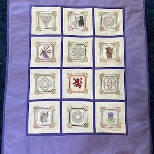 May include: A lavender quilt with embroidered squares displaying Celtic knots, animals, and a thistle. The squares are framed with gold trim on a white background. The quilt has a lavender border.