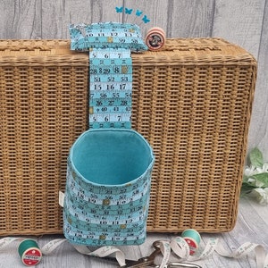 May include: A blue fabric storage container with a measuring tape design hanging on a wicker basket. The container is filled with sewing supplies, including spools of thread and a pincushion.