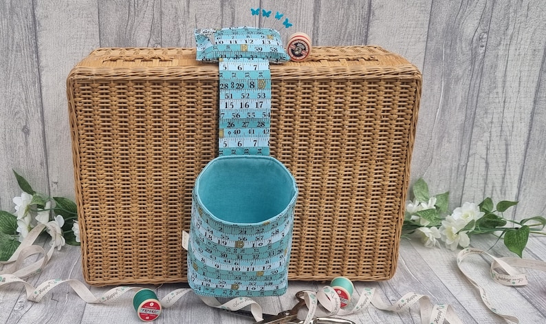May include: A blue fabric storage container with a measuring tape design hanging on a wicker basket. The container is filled with sewing supplies, including spools of thread and a pincushion.