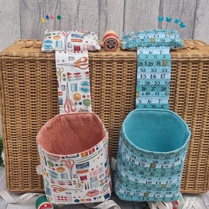 May include: Two fabric hanging organisers with a sewing theme. The organiser on the left is peach coloured with a white background and a pattern of sewing tools. The organiser on the right is teal blue with a white background and a pattern of measuring tape. Both organisers are hanging from a wicker basket.