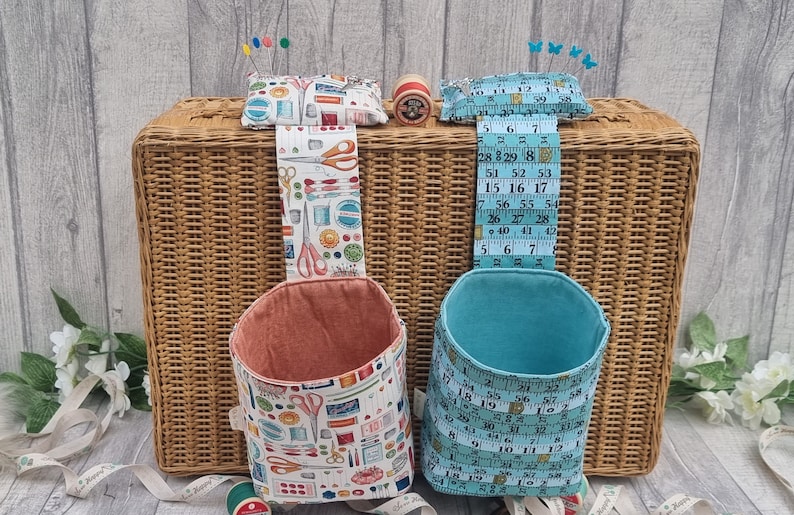 May include: Two fabric hanging organisers with a sewing theme. The organiser on the left is peach coloured with a white background and a pattern of sewing tools. The organiser on the right is teal blue with a white background and a pattern of measuring tape. Both organisers are hanging from a wicker basket.