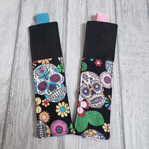 Könnte beinhalten: Zwei schwarze Stoff-Lesezeichenhalter mit bunten Zucker Schädel Designs. Die Lesezeichenhalter haben eine schwarze Stoffrückseite und ein blaues und rosa Band.