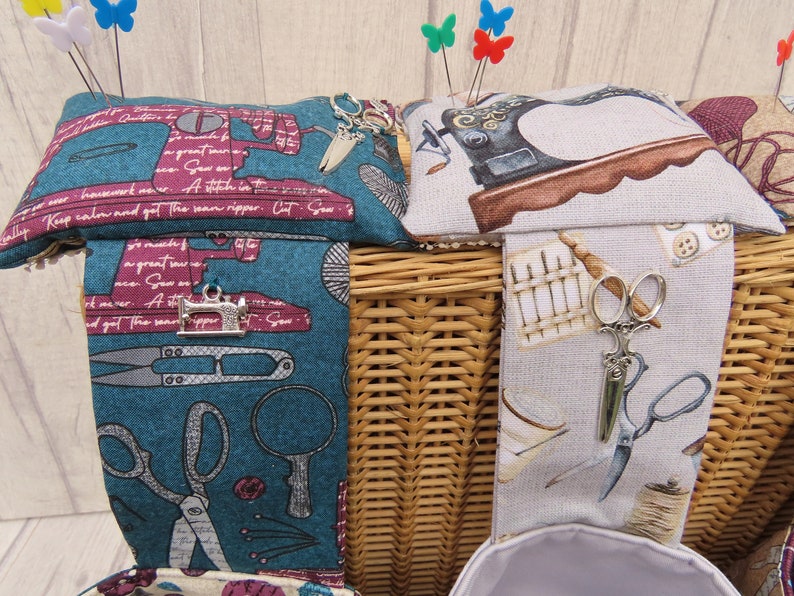 May include: Two wicker baskets with fabric lids. The lids are decorated with sewing-themed patterns and have pincushions on top. One lid has a teal background with a sewing machine, scissors, and thread. The other lid has a beige background with a sewing machine, buttons, and a spool of thread. Both lids have a pincushion with colourful pins.