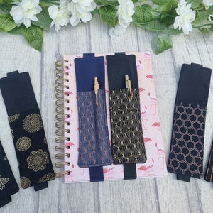May include: Six black fabric pen holders with different gold and copper patterned designs. The holders are attached to a notebook with a pink flamingo pattern.