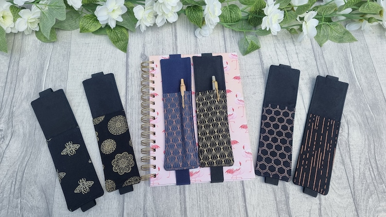 May include: Six black fabric pen holders with different gold and copper patterned designs. The holders are attached to a notebook with a pink flamingo pattern.