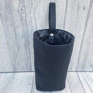 Puede incluir: Una bolsa de tela negra con un cord&oacute;n de ajuste negro y una correa negra. La bolsa est&aacute; colgada de una superficie de madera.