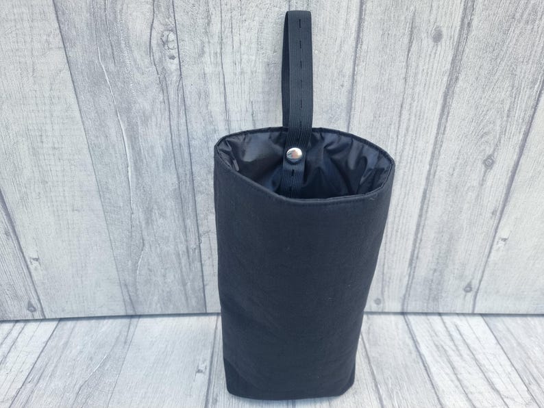 Puede incluir: Una bolsa de tela negra con un cord&oacute;n de ajuste negro y una correa negra. La bolsa est&aacute; colgada de una superficie de madera.