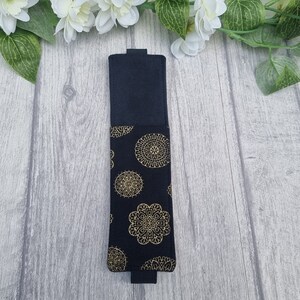 May include: A black fabric pencil case with a gold mandala pattern. The case is closed and lying flat on a light wood surface.