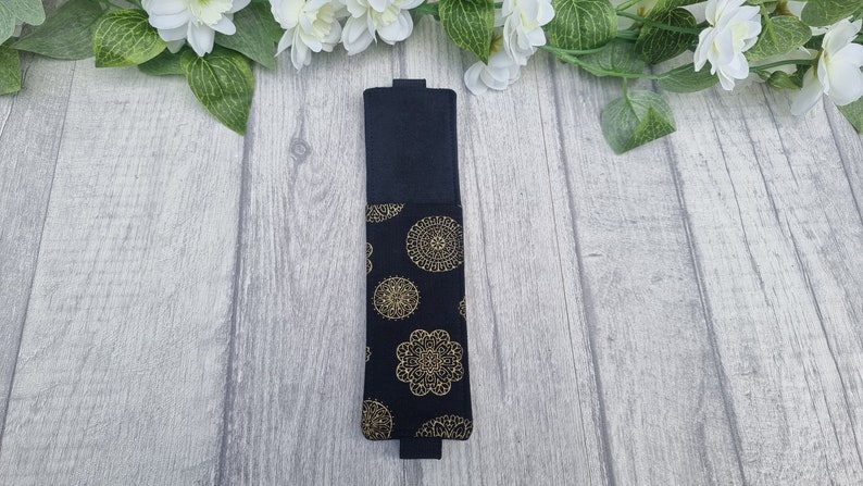 May include: A black fabric pencil case with a gold mandala pattern. The case is closed and lying flat on a light wood surface.