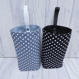 Puede incluir: Dos bolsas de almacenamiento de tela colgantes con lunares blancos sobre un fondo gris y negro. Las bolsas tienen correas blancas y negras.