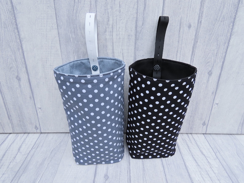 Puede incluir: Dos bolsas de almacenamiento de tela colgantes con lunares blancos sobre un fondo gris y negro. Las bolsas tienen correas blancas y negras.