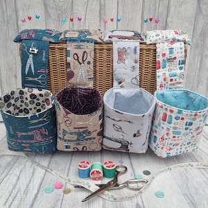 Può includere: Sei cesti di stoffa per la conservazione con stampe a tema cucito, tra cui forbici, bottoni e macchine da cucire. I cesti sono appesi a un cesto di vimini con un cuscino per spilli in cima. Ci sono anche bobine di filo e forbici sul tavolo.