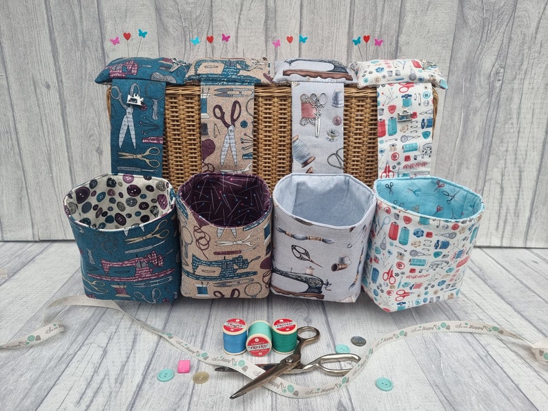 May include: Six fabric storage baskets with sewing-themed prints, including scissors, buttons, and sewing machines. The baskets are hanging on a wicker basket with a pincushion on top. There are also spools of thread and scissors on the table.