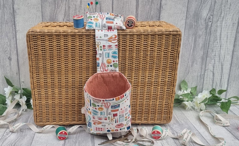 May include: A white fabric hanging organiser with a pink lining and a colourful print of sewing tools. The organiser is hanging from a wicker basket with a spool of thread and a pincushion on top.