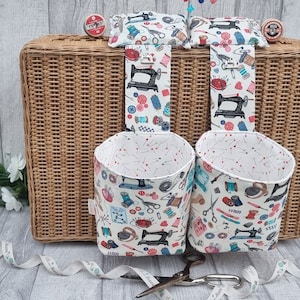 May include: Two white fabric hanging storage baskets with a sewing machine and other sewing supplies pattern. The baskets are hanging from a wicker suitcase.