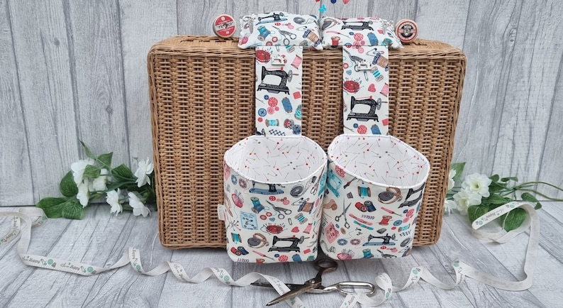 May include: Two white fabric hanging storage baskets with a sewing machine and other sewing supplies pattern. The baskets are hanging from a wicker suitcase.
