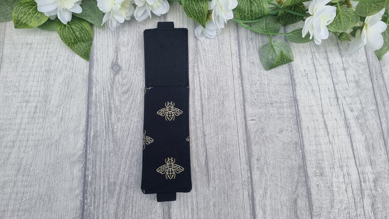 May include: Black fabric pencil case with a gold bee pattern. The case is closed and has a flap.