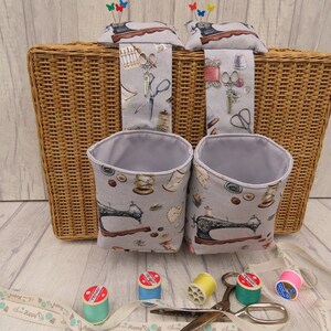May include: Two grey fabric hanging storage containers with a sewing machine print. The containers are hanging on a wicker basket. There are spools of thread and scissors on a wooden surface.